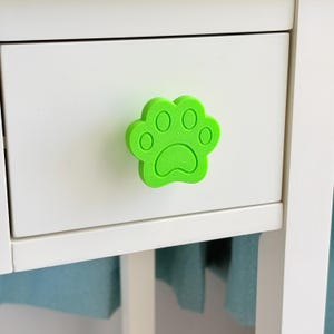 May include: A bright green paw print shaped drawer pull on a white drawer. The drawer is part of a white cabinet. The paw print has five circular pads and a curved bottom pad. The cabinet has a light blue fabric hanging below.