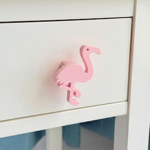 May include: A light pink flamingo-shaped drawer knob attached to a plain white drawer front. The knob features a stylized silhouette of a standing flamingo with a curved neck, a pointed beak, and a single small circular eye detail in a matte finish.