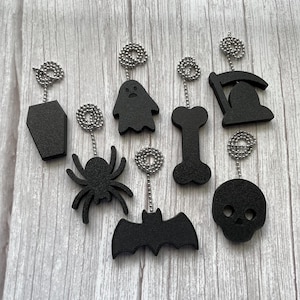 May include: A collection of black Halloween-themed charms, including a coffin, ghost, bone, grim reaper, spider, bat, and skull. Each charm is attached to a silver ball chain, perfect for jewelry or craft projects. The charms are displayed on a light wood-grain surface.