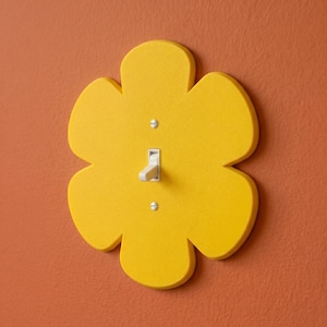 May include: A yellow flower-shaped light switch cover with a standard toggle switch. The cover is mounted on an orange wall. The switch is in the 'on' position. The design adds a decorative touch to a functional item.