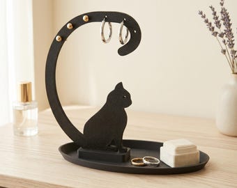 May include: Black cat-shaped jewelry organizer with a curved top for hanging earrings. The organizer has a tray base for rings and small jewelry items. Gold-colored studs adorn the curved top. A small white box and two rings are on the tray.