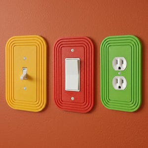 May include: Three colorful electrical outlet covers in yellow, red, and green. The yellow cover has a toggle switch, the red cover has a rocker switch, and the green cover has two electrical outlets. All covers have a grooved, rectangular design.