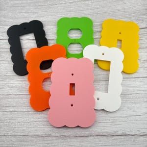 May include: A collection of colorful, scalloped-edge light switch and outlet covers in black, orange, green, yellow, pink, and white. Each cover has a different opening for various electrical configurations. The covers are made of a solid material.