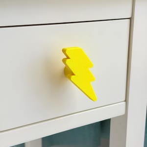 May include: A bright yellow lightning bolt-shaped drawer pull is mounted on a white drawer. The drawer pull has a textured surface and is a vibrant accent against the white background. This decorative hardware adds a playful touch to furniture.