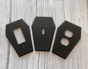 May include: Three black coffin-shaped wall plates for light switches and outlets. The plates have a textured finish and are designed to look like a coffin.