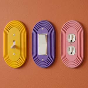 May include: Three colorful electrical wall plates: yellow light switch, purple light switch, and pink electrical outlet. Each plate has a ridged, oval design. The light switches are white, and the electrical outlet has two slots and a grounding hole.