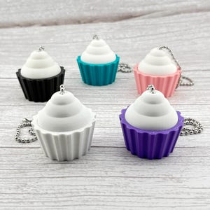 May include: Five cupcake-shaped keychains in black, teal, pink, white, and purple. Each has a white frosting top and a silver ball chain. The cupcake bases are ribbed.