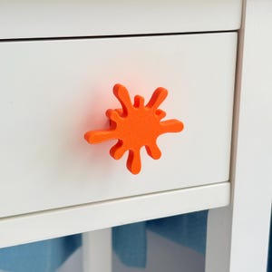 May include: An orange, splash-shaped drawer pull is mounted on a white drawer. The drawer pull has a textured surface and is a bright, eye-catching accent. The drawer is part of a white cabinet or dresser.