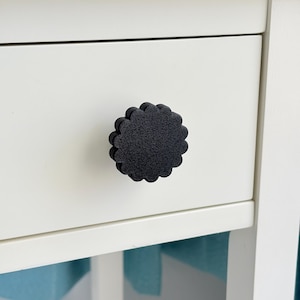 May include: Close-up of a white drawer with a black, flower-shaped knob. The knob has a textured surface and is centered on the drawer front. The drawer is part of a white cabinet or dresser.