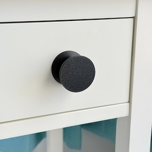 May include: Close-up of a white drawer with a black, textured, round knob. The knob is centered on the drawer, providing a simple, modern aesthetic. The drawer is part of a white cabinet.