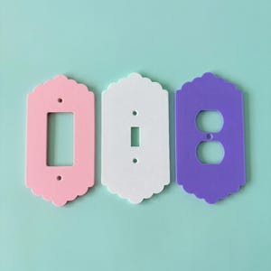 May include: Three decorative light switch covers in pink, white, and purple. Each cover has a scalloped edge and a different opening: a rectangular opening, a single switch opening, and a double outlet opening.