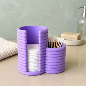 May include: A lavender-colored bathroom organizer with a ribbed design. It features a section for cotton pads and a smaller compartment holding cotton swabs. The organizer sits on a light-colored surface, with a soap dish and bottle in the background.