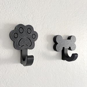 May include: Two black wall hooks. One hook is shaped like a dog paw print, and the other is shaped like a dog bone. Both hooks are made of a matte black material and are mounted on a white wall.