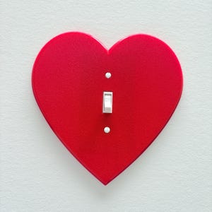 May include: A red heart-shaped light switch cover with a white toggle switch and two silver screws. The cover is mounted on a white wall, creating a striking contrast. The design is simple yet eye-catching.