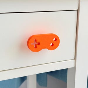 May include: An orange, 3D-printed drawer pull in the shape of a classic video game controller. The controller features a directional pad and two circular buttons. It is mounted on a white drawer.