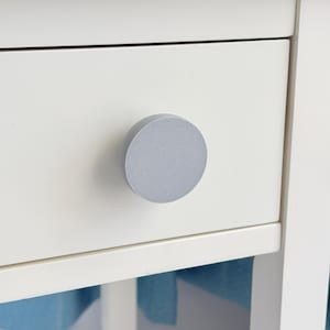 May include: Close-up of a white drawer with a round, light gray knob. The knob is centered on the drawer front, providing a simple, modern aesthetic. The drawer is part of a larger white furniture piece.