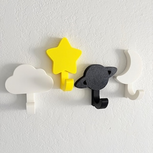 May include: Four decorative wall hooks in the shape of a cloud, star, planet, and moon. The cloud and moon are white, the star is yellow, and the planet is black. Each hook has a curved bottom for hanging items.