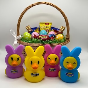 Könnte beinhalten: Ein Osterkorb gefüllt mit Leckereien, darunter eine Tüte Peeps-Süßigkeiten, Schokoladenhasen und dekorierte Eier. Davor befinden sich vier bunte Peeps-Entenspielzeuge, die jeweils eine Hasenkapuze tragen. Die Spielzeuge sind lila, gelb, rosa und blau.