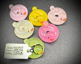 May include: A collection of colorful, duck-shaped donut charms. The charms are in shades of pink, yellow, green, and clear, each with a small duck head and orange beak. The text "Grab Yours Today" is on a white tag.