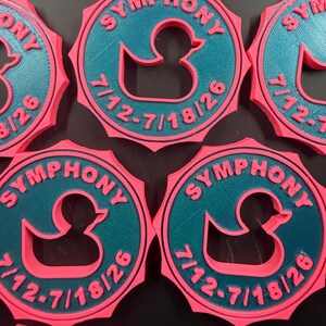 May include: Five circular pink and teal plastic medallions featuring a central cutout of a rubber duck. The text SYMPHONY is embossed in pink at the top, and the date 7/12-7/18/26 is embossed in pink at the bottom of each teal circular center.