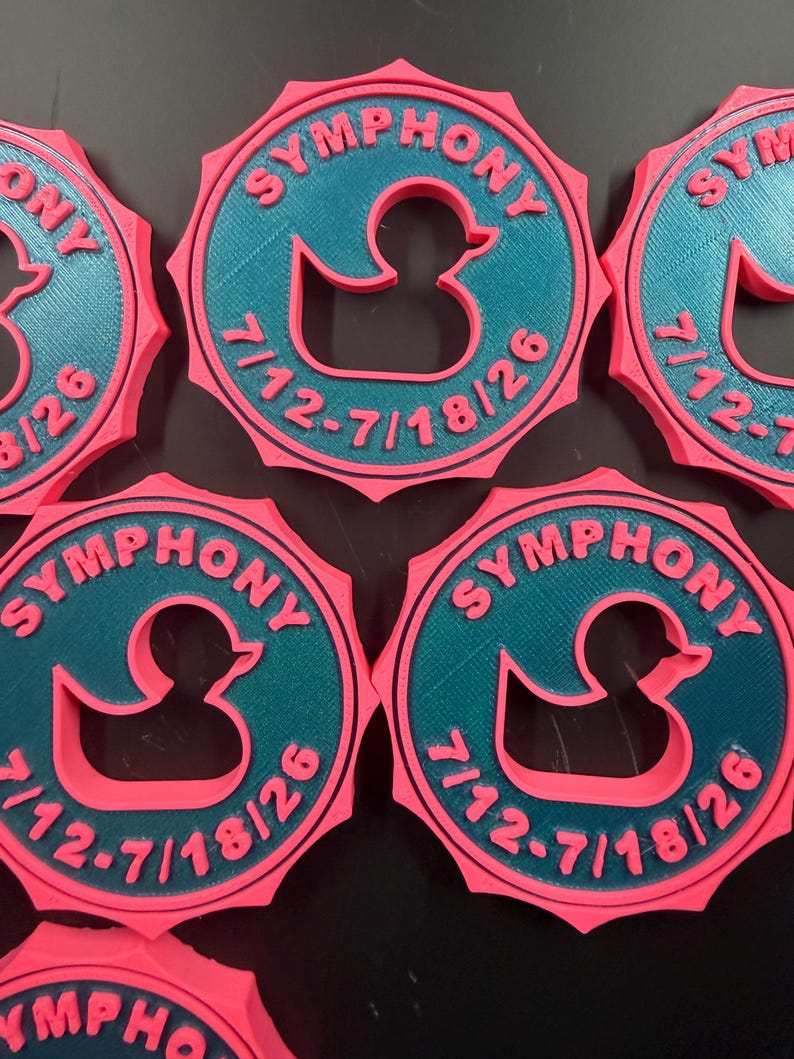 May include: Five circular pink and teal plastic medallions featuring a central cutout of a rubber duck. The text SYMPHONY is embossed in pink at the top, and the date 7/12-7/18/26 is embossed in pink at the bottom of each teal circular center.