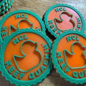 May include: A set of circular orange coasters with a teal scalloped border. Each coaster features a cutout of a rubber duck in the center and the raised teal text NCL KEEP QUACKIN. The coasters have a textured surface and a layered design.