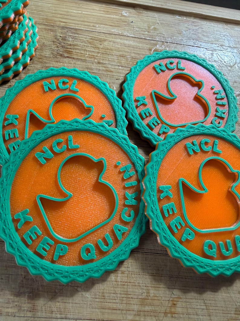 May include: A set of circular orange coasters with a teal scalloped border. Each coaster features a cutout of a rubber duck in the center and the raised teal text NCL KEEP QUACKIN. The coasters have a textured surface and a layered design.