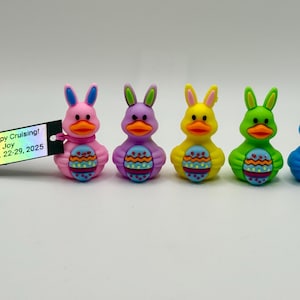 May include: Five brightly coloured rubber duck figurines, each designed as a duck-bunny hybrid, holding an Easter egg. The ducks are pink, purple, yellow, green, and blue. A tag with text is on the left.
