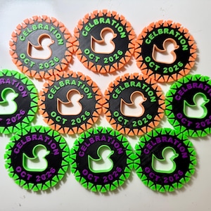 May include: Ten circular black plastic tags with a serrated orange or green border. Each tag features a cutout of a rubber duck in the center, with the text CELBRATION and OCT 2026 embossed in orange or purple lettering around the duck silhouette.