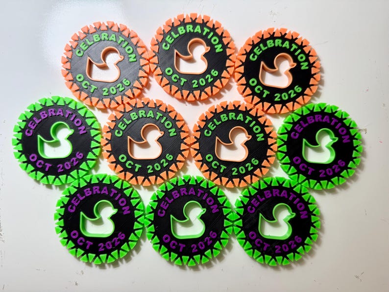 May include: Ten circular black plastic tags with a serrated orange or green border. Each tag features a cutout of a rubber duck in the center, with the text CELBRATION and OCT 2026 embossed in orange or purple lettering around the duck silhouette.