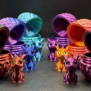 May include: A selection of vibrant, 3D-printed dragon figures and coordinating lidded pots. The dragons are in shades of blue, orange, purple, and red, with large eyes. The pots feature a textured, scale-like design. The scene is set against a dark background.
