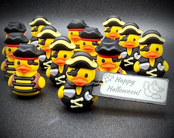 Puede incluir: Un grupo de patitos de goma amarillos disfrazados de piratas, con sombreros y chalecos negros, y parches rojos en los ojos. Un pato sostiene un letrero con la frase "Happy Halloween!" en blanco, con un dibujo de un murciélago y una telaraña.