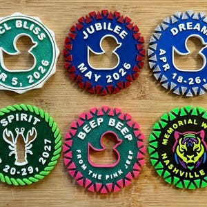 May include: Six circular 3D-printed cruise ship tags featuring various icons like ducks, a lobster, and a tiger. Each tag displays specific text including NCL BLISS APR 5 2026, JUBILEE MAY 2026, DREAM APR 18-26 2026, SPIRIT MAR 20-29 2027, BEEP BEEP FROM THE PINK JEEF, and MEMORIAL WKND NASHVILLE 2026.