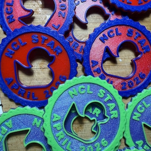 May include: A collection of circular, gear-shaped plastic tags featuring a central duck silhouette cutout. Each tag is embossed with the text NCL STAR and APRIL 2026. The tags are available in red with blue borders or bright green with blue borders.
