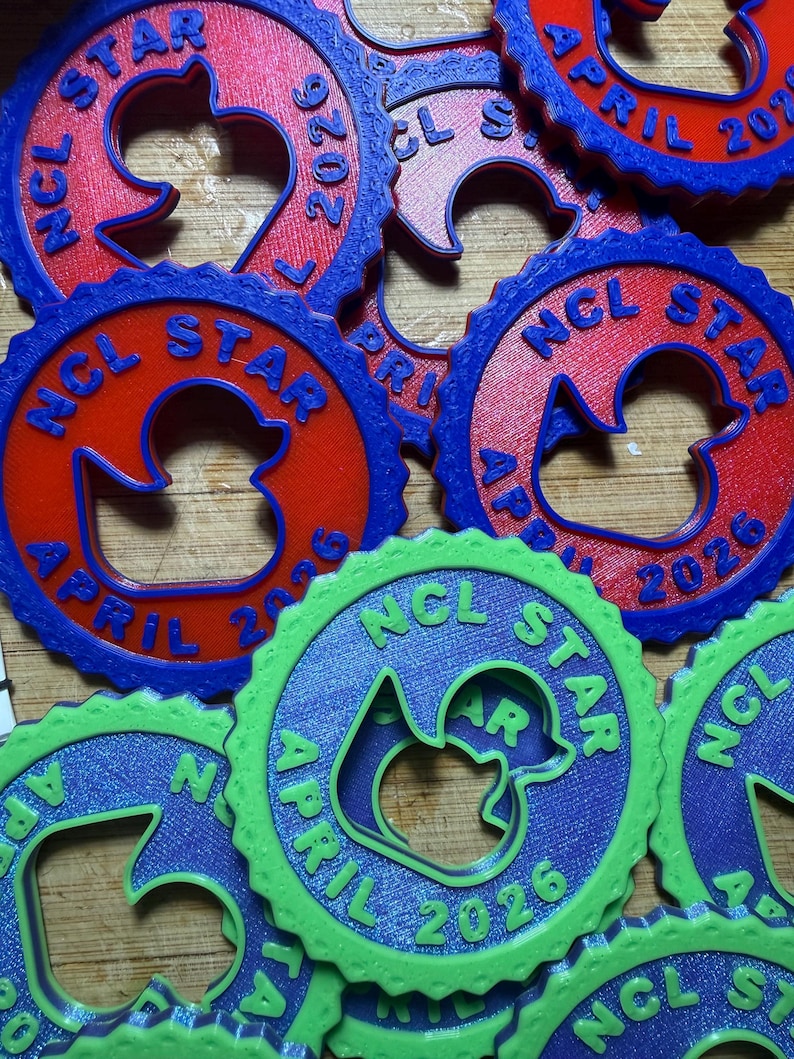 May include: A collection of circular, gear-shaped plastic tags featuring a central duck silhouette cutout. Each tag is embossed with the text NCL STAR and APRIL 2026. The tags are available in red with blue borders or bright green with blue borders.