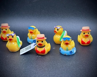 May include: A collection of yellow rubber duck figurines, some wearing sunglasses, hats, and swimsuits. One duck holds a surfboard. Some ducks are in inflatable rings. A tag is visible.