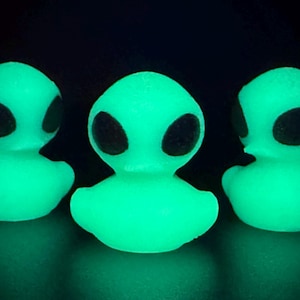 May include: Three glowing green alien-shaped figures with large black oval eyes. The figures are duck-like in shape, with a rounded body and head. The background is black.