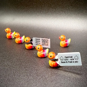 May include: A collection of small, colorful duck figurines with orange, yellow, and rainbow-colored details. Each duck has a tag attached with text and a QR code. The tags read "Quack Quack Post a Photo" and "Carnival 3/31-4/4 Scan & Post a pic!"