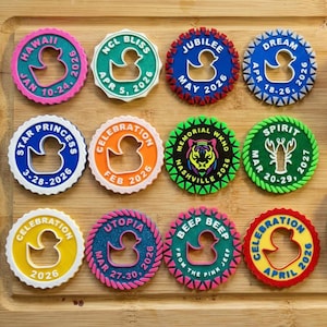 May include: A collection of colorful, circular decorations with a duck silhouette cutout. Each decoration features text indicating a location and date, such as "Hawaii Jan 10-24 2026" and "NCL Bliss Apr 5, 2026". The decorations are arranged on a wooden surface.