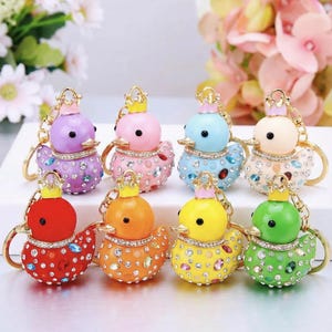 Pode incluir: Oito chaveiros de pato adornados com joias, cada um com uma coroa dourada e strass brilhantes. Os patos são em tons de roxo, rosa, azul, creme, vermelho, laranja, amarelo e verde. Dispostos em duas fileiras.