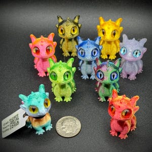 May include: A selection of brightly coloured 3D-printed dragon figurines. The dragons are in shades of pink, green, blue, and yellow. A coin is included for scale. Each dragon has large eyes and a unique colour scheme. One dragon has a tag attached.