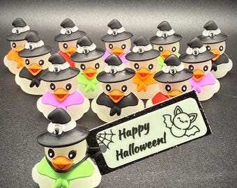 May include: A collection of Halloween-themed rubber duckies, each wearing a black witch hat and a colorful scarf. The ducks are arranged in a group, with a tag that reads "Happy Halloween!" and a bat illustration.