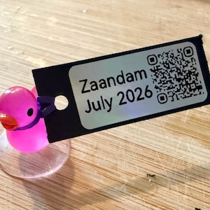 Puede incluir: Un patito de goma rosa con una banda morada en el cuello y una base transparente. Una etiqueta negra con el texto "Zaandam July 2026" y un código QR.