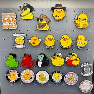 May include: A collection of yellow duck-shaped pins and badges with various designs. Some ducks wear hats and sunglasses, while others hold knives or have text like "Choose Love" and "Bad Bitch Club".