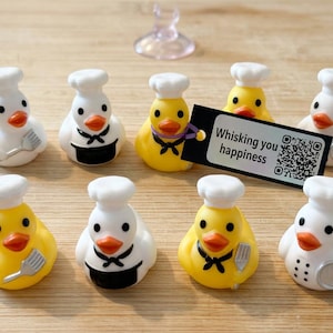 May include: A collection of chef duck figurines, some yellow and some white, each wearing a chef's hat and apron. Some ducks hold a spatula. A tag reads "Whisking you happiness" with a QR code.