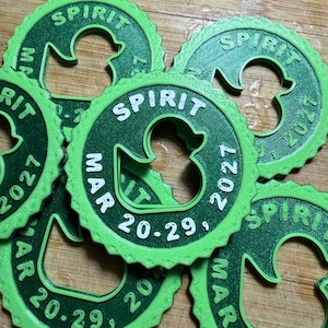 May include: A collection of circular green plastic tags with a scalloped edge, each featuring a central cutout in the shape of a duck. The text "SPIRIT" is embossed at the top, with "MAR 20-29, 2027" printed along the bottom curve of each tag.
