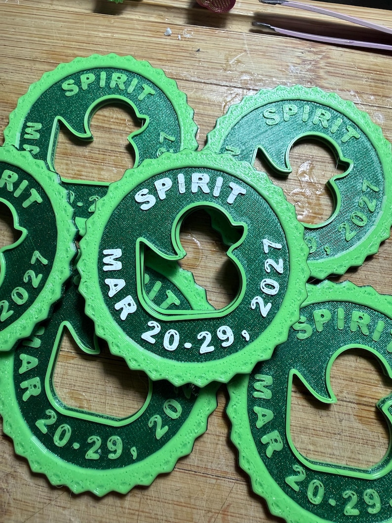 May include: A collection of circular green plastic tags with a scalloped edge, each featuring a central cutout in the shape of a duck. The text "SPIRIT" is embossed at the top, with "MAR 20-29, 2027" printed along the bottom curve of each tag.