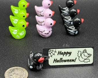 May include: A collection of Halloween-themed rubber duckies in green, lavender, and black. The green ducks have ghost and dot designs. The lavender ducks have bat wing designs. The black ducks have skeleton designs. A tag reads "Happy Halloween!"