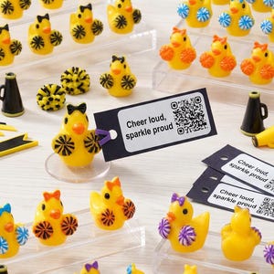 Könnte beinhalten: Eine Ansammlung gelber Entenfiguren mit Schleifen und Pompons, angeordnet auf transparenten Präsentationsständern. Cheerleader-Zubehör wie Megaphone und Bänder sind verstreut. Der Text "Cheer loud, sparkle proud" ist auf Etiketten zu sehen.