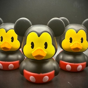 Può includere: Tre paperelle di gomma a forma di Topolino. Le paperelle hanno corpi neri, facce gialle, becchi arancioni e fondi rossi con cerchi bianchi. Le orecchie delle paperelle sono nere e rotonde.